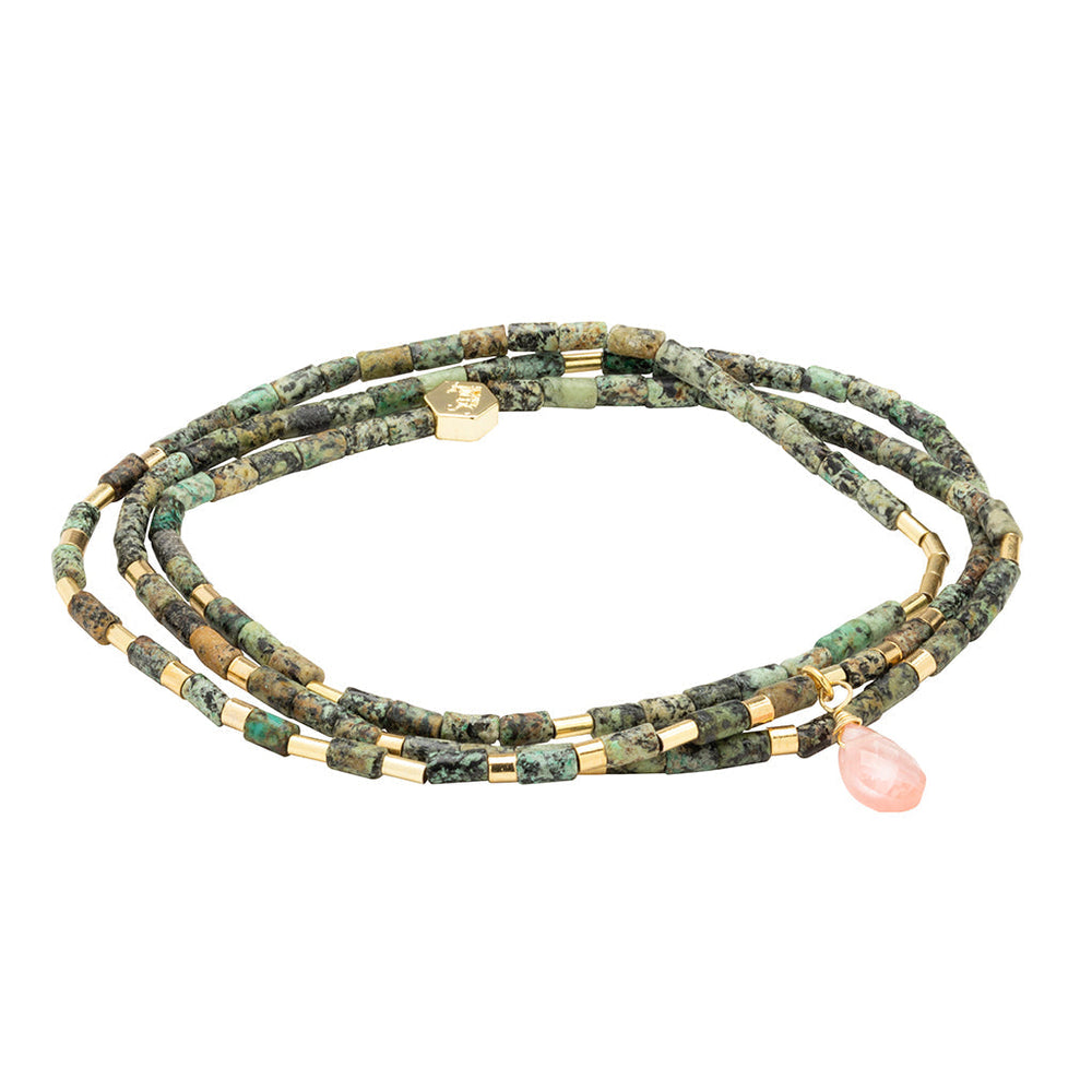 The Transformation African Turq Gold Teardrop Wrap by Scout Jewelry features green and gold cylindrical beads with a pink teardrop charm, coiled in a loose spiral on a white background.
