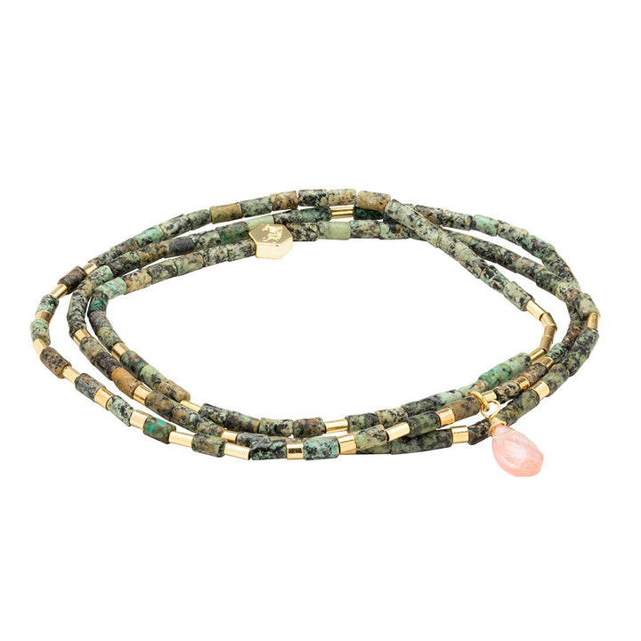 The Transformation African Turq Gold Teardrop Wrap by Scout Jewelry features green and gold cylindrical beads with a pink teardrop charm, coiled in a loose spiral on a white background.