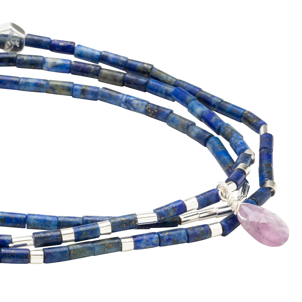 A close-up of the Scout Jewelry Clarity Lapis Silver Teardrop Wrap, showcasing cylindrical blue and silver beads with a faceted amethyst teardrop pendant wrapped in silver wire.