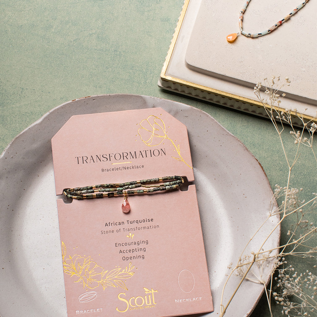 The Transformation African Turq Gold Teardrop Wrap by Scout Jewelry is displayed on a pink “TRANSFORMATION” card atop a ceramic plate, with a matching necklace placed nearby on a beige tray accented with dried flowers.