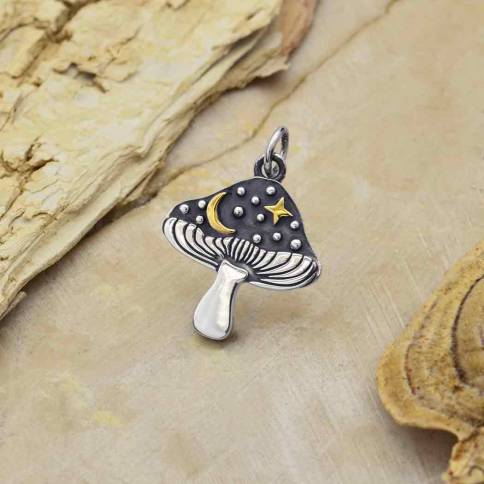 The Silver Mushroom Charm with Bronze Star and Moon by Nina Designs features a dark cap, gold crescent moon, bronze star and moon, and silver dots, displayed on a textured beige surface with pieces of wood nearby.