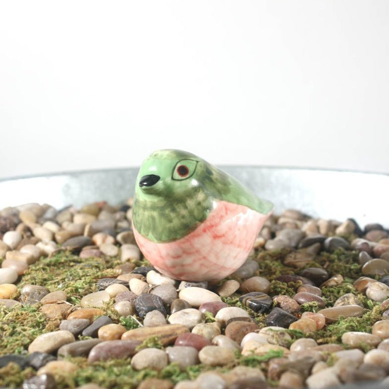 The CreativeCoOp Painted Stoneware Bird, a small hand-painted ceramic figurine in green and pink hues, sits whimsically wide-eyed on moss and assorted pebbles within a metal tray.