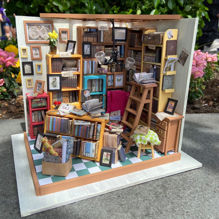 The Sam's Study Room DIY Miniature Kit by Hands Craft is a detailed 3D puzzle of a cozy bookstore, featuring tiny books, shelves, a ladder, reading chair, table with checkered cloth, decorative plants, and bright flowers in the background.
