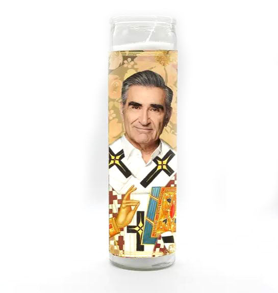 The JohnnyRose Candle by BOBBYK BOUTIQUE is an unscented decorative piece featuring an illustration of a man in a white shirt, surrounded by religious and ornate patterns, along with tall geometric shapes and a floral background.