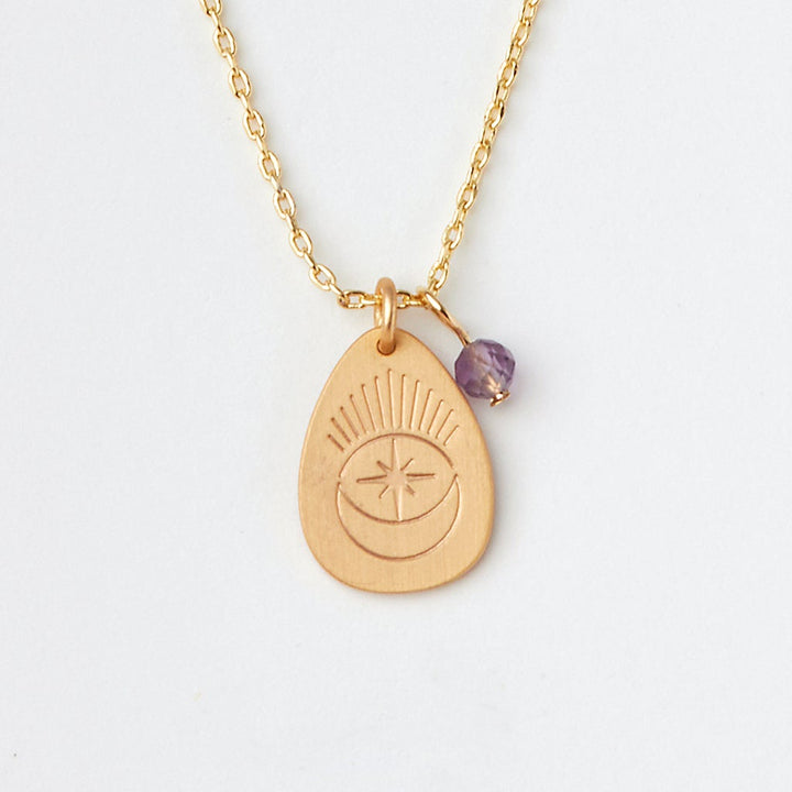 Introducing the "Intention Neck Amethyst/Gold" by Scout Jewelry: a gold necklace featuring an engraved teardrop-shaped pendant with a crescent moon and starburst design. The pendant includes a small purple semi-precious amethyst stone, all elegantly suspended from a fine, adjustable brass chain. Displayed beautifully against a plain white background.