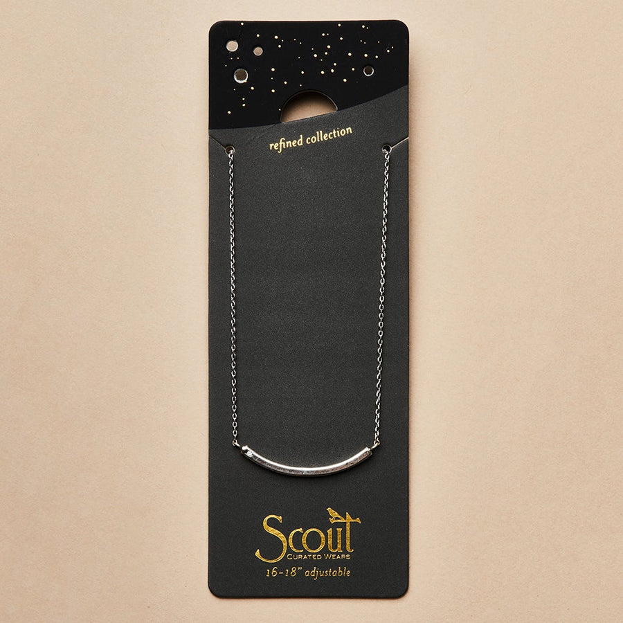 The "Refined Neck Comet/Silver" by Scout Jewelry is a silver necklace featuring a curved bar design with CZ stones. It comes in black and gold packaging with a starry pattern, labeled "Scout Curated Wears," set against a soft beige background displaying the text "refined collection.