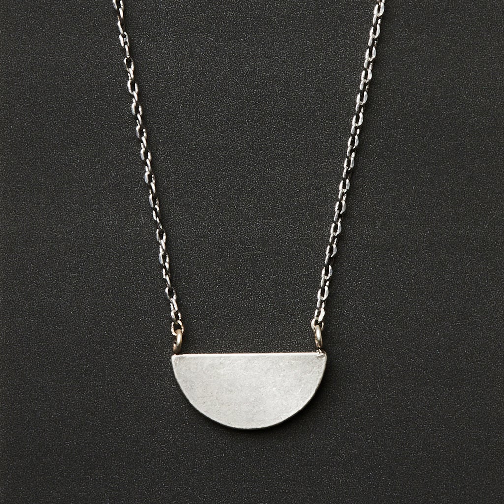 The Refined Neck Half Moon/Silver by Scout Jewelry features a simple adjustable silver chain with a flat, matte half-circle pendant at the center, displayed against a black background.
