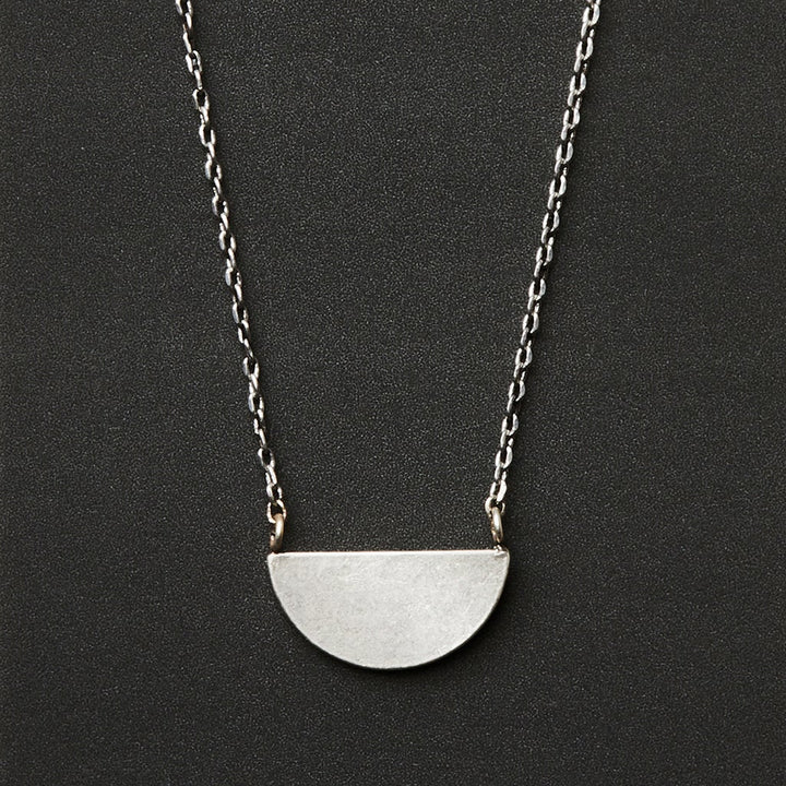 The Refined Neck Half Moon/Silver by Scout Jewelry features a simple adjustable silver chain with a flat, matte half-circle pendant at the center, displayed against a black background.