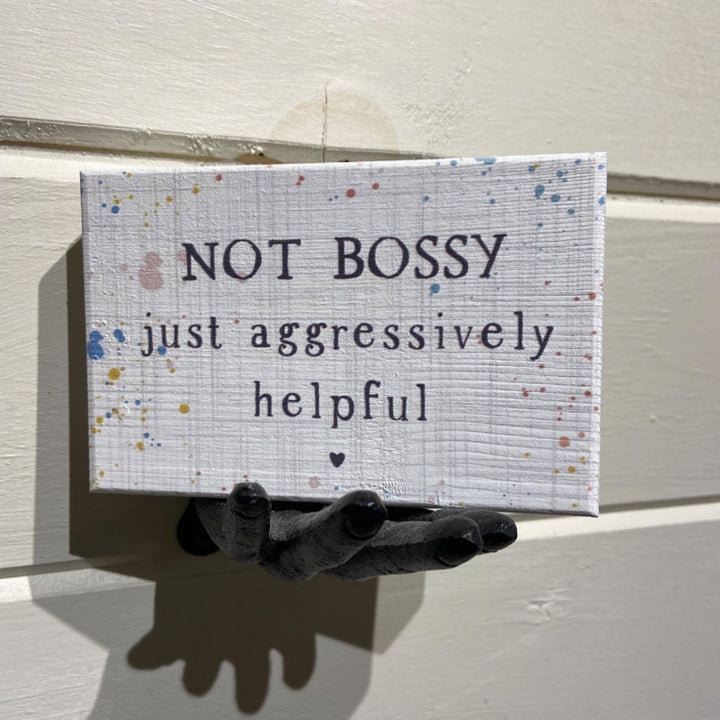 A small, handcrafted sign named "Not Bossy" by SincereSurroundings features a speckled pattern and the words "NOT BOSSY just aggressively helpful." It is elegantly displayed on a wall-mounted decorative hand for easy showcasing.