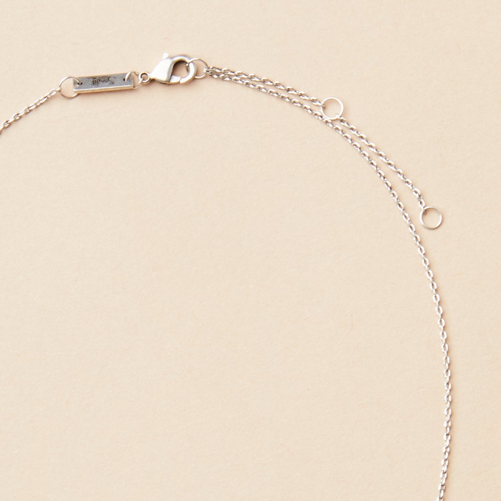 A piece from Scout Jewelry, the Refined Neck Crescent in silver, elegantly arranged in a partial circle on a plain beige background. This delicate necklace features a lobster clasp and a small rectangular tag near the clasp that highlights its exquisite craftsmanship.