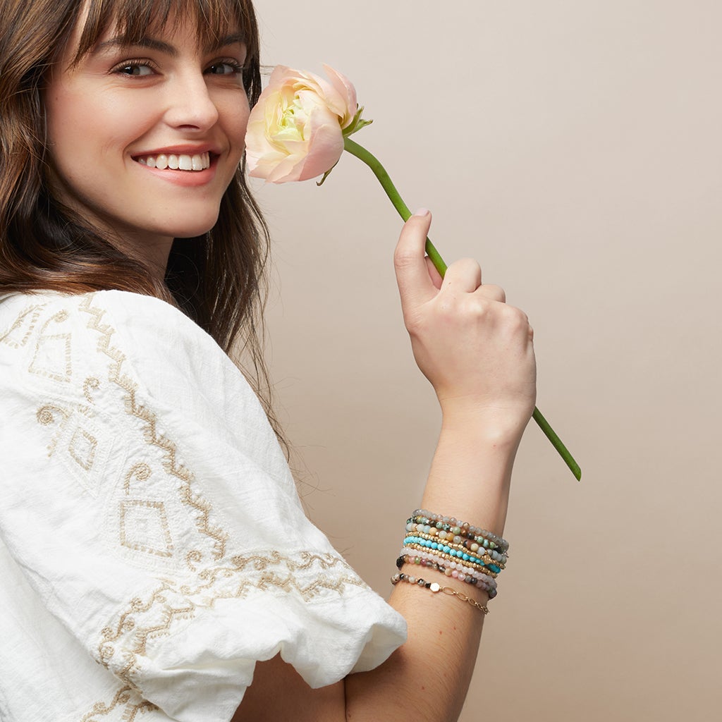 A woman with long brown hair smiles while holding a peach-colored flower against a light beige background. She is wearing a white embroidered top and multiple Mini Bracelet Turquoise/Gold bracelets from Scout Jewelry on her wrist, featuring faceted stone beads on stretch cord.