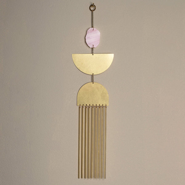 The Nebula Rose Quartz Wall Hanging by Scout Jewelry features a pink oval stone, stacked brass semicircles, and vertical brass rod fringe, all suspended from a chain against a plain beige wall.