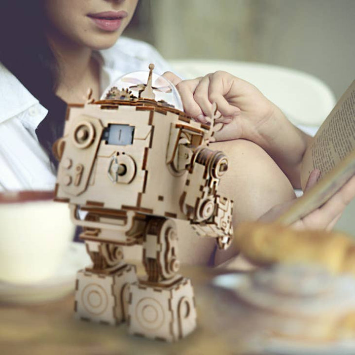 A woman reads a book while sitting at a table with a cup and pastries. In the foreground, a DIY 3D Steam Punk Music Box: Orpheus from Hands Craft stands, capturing the focus with its intricate steampunk design. The scene is cozy and relaxed.