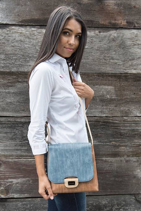 A woman with long dark hair, in a white button-up shirt and jeans, stands before a wooden wall holding the Liz Soto Handbags Amy Crossbody - Denim, featuring an adjustable strap and buckle clasp.