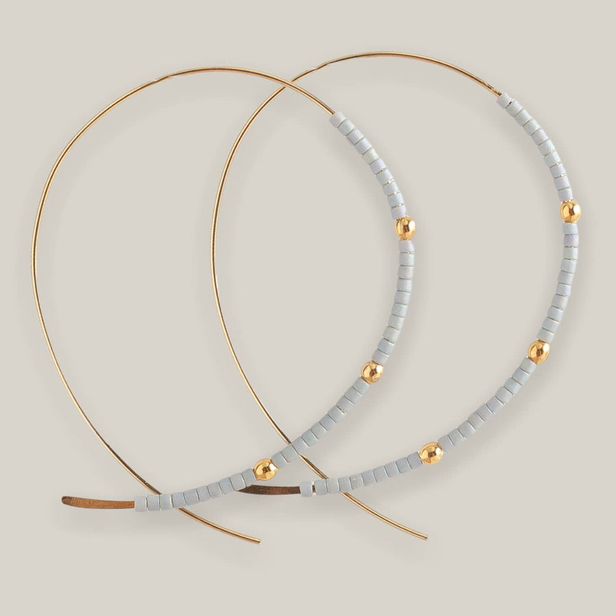 The Confetti Earrings, Grey by Lenny & Eva feature large gold-plated hoops with pale gray Miyuki beads and spaced gold accents, shown against a neutral beige background.