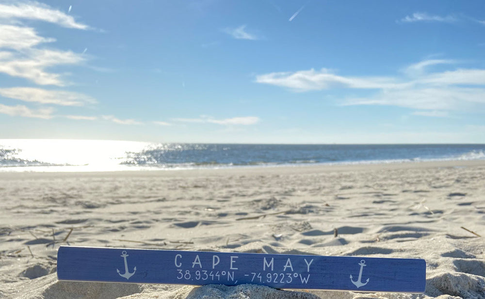 A sandy beach with a calm ocean under a clear blue sky shows the "Cape May Coordinates" plank by SincereSurroundings, featuring anchor symbols and the text "CAPE MAY 38.9344°N, -74.9223°W," proudly displayed on the sand like a unique shelf treasure.