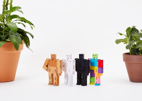 Four brightly colored wooden toy robots are arranged in a line between two potted plants. Crafted from a combination of wood and plastic, these figures feature articulated limbs and each boasts a unique color scheme. Among them, the Cubebot Micro Yellow Multi by Areaware introduces an eye-catching burst of vibrant yellow against the plain white backdrop.