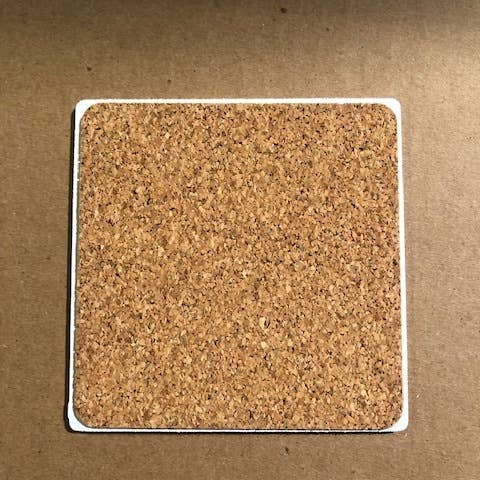 A square cork coaster with rounded edges on a brown background, named the Internet Stopped coaster by Drinks On Me. This minimalist piece features a natural cork texture and remains simple and unadorned.