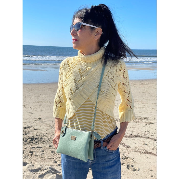 A woman with dark hair in a ponytail, wearing sunglasses, a yellow knit sweater over a striped top, blue jeans, and the Ella Crossbody - Almond by Liz Soto Handbags stands on a sandy beach with the ocean and blue sky in the background.