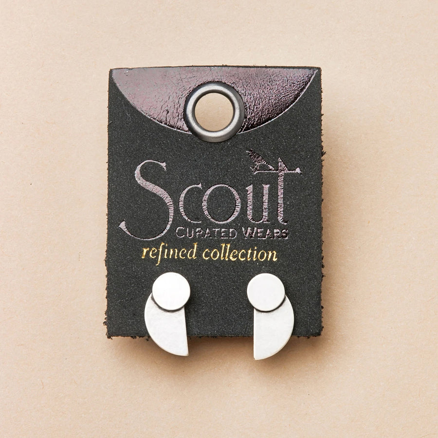 A pair of Eclipse Stud Silver earrings by Scout Jewelry, crafted from 925 sterling silver with a geometric design, are displayed on a black leather card labeled "Scout Curated Wears Refined Earring Collection" and accented with burgundy and a circular hole.