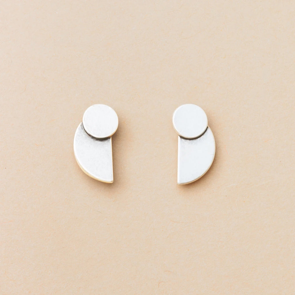The Eclipse Stud Silver earrings by Scout Jewelry, crafted from 925 sterling silver, feature a small circle joined to a larger geometric shape and are shown on a light beige background as part of the Refined Earring Collection.