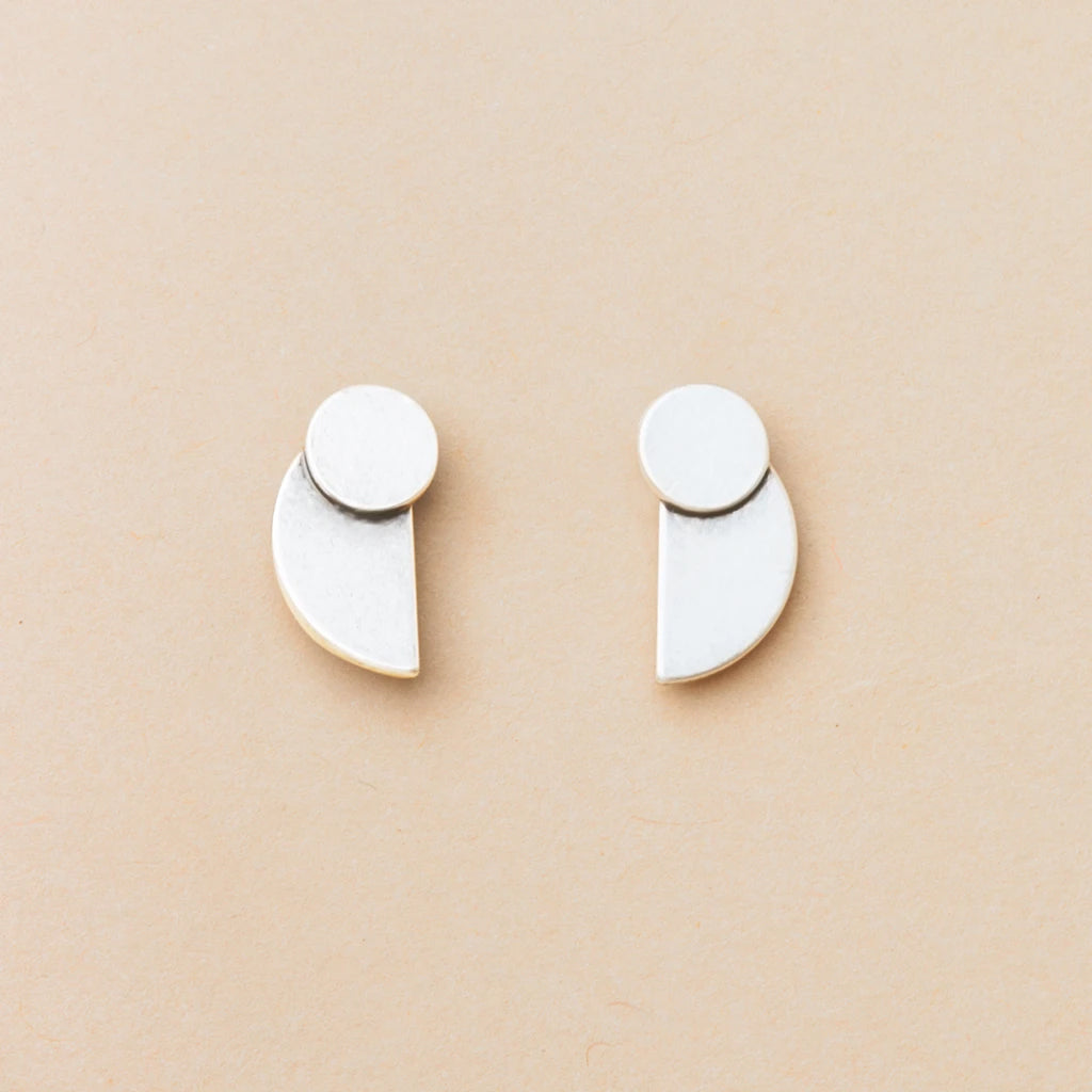 The Eclipse Stud Silver earrings by Scout Jewelry, crafted from 925 sterling silver, feature a small circle joined to a larger geometric shape and are shown on a light beige background as part of the Refined Earring Collection.