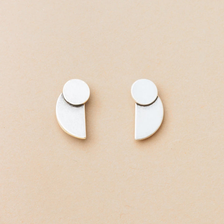 The Eclipse Stud Silver earrings by Scout Jewelry, crafted from 925 sterling silver, feature a small circle joined to a larger geometric shape and are shown on a light beige background as part of the Refined Earring Collection.