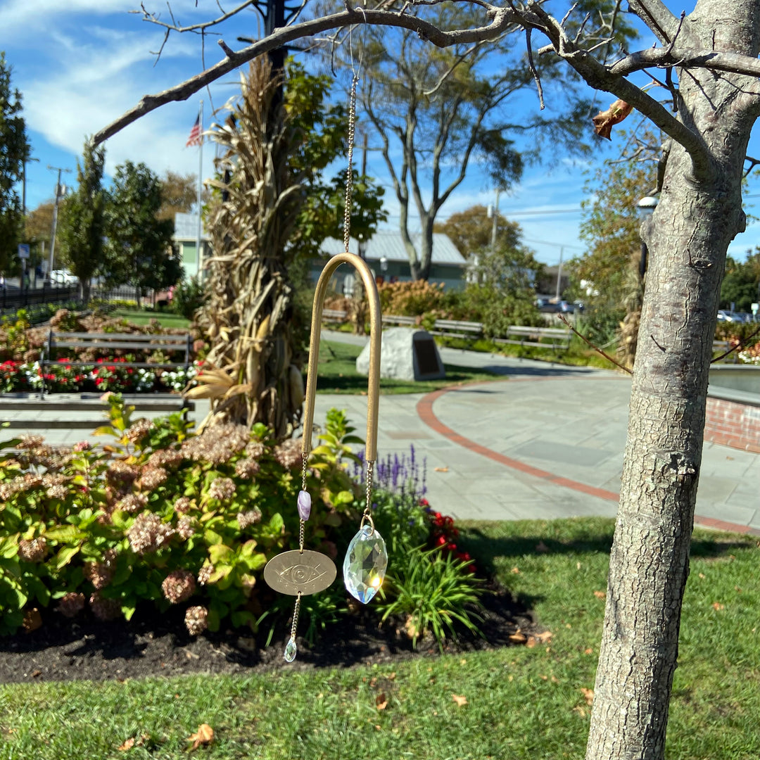 The Scout Jewelry Evil Eye Amethyst Suncatcher hangs from a tree branch, creating rainbows in the sunlight. A park with colorful flowers and trees under a clear blue sky serves as the backdrop.