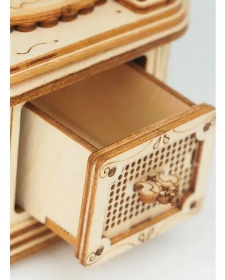 A close-up of a small, intricately designed drawer partially open on the Hands Craft 3D Wooden Puzzle Gramophone. The drawer front features a detailed pattern with a small handle, reminiscent of modern vintage style. The light-colored wood has darker accents along the edges and carved decorations, much like those found on a fine gramophone detail.