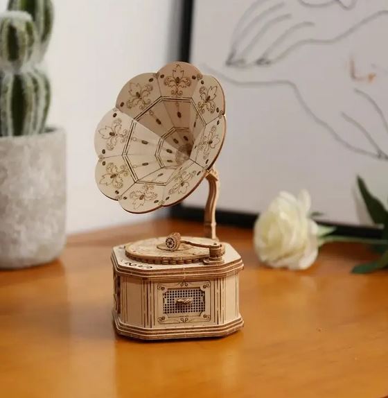 A 3D Wooden Puzzle Gramophone from Hands Craft sits on a wooden surface, embodying a modern vintage style. The gramophone features a detailed floral design on the horn. In the background, a potted cactus and a framed artwork are visible, while a white rose lies next to the gramophone.