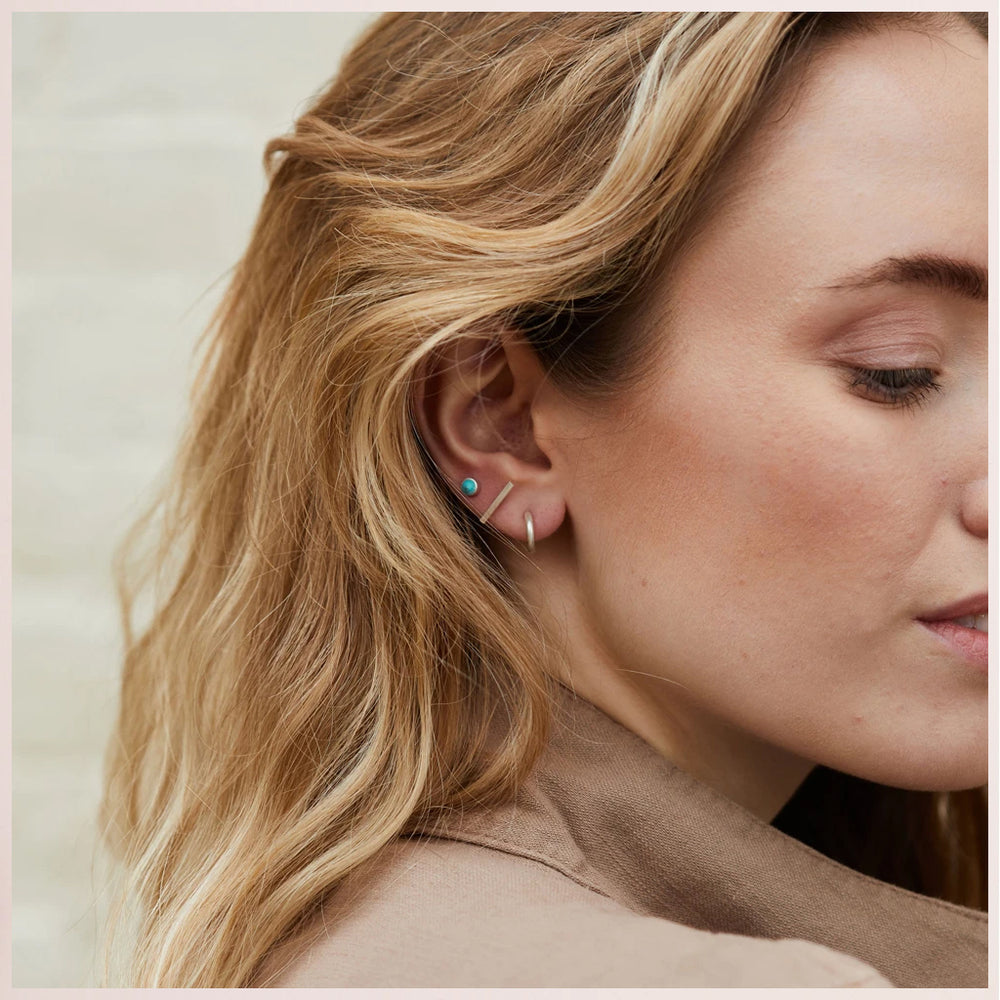 A woman with wavy blonde hair wears Scout Jewelry's Scarlett Stud Trio Turquoise Silver—featuring a semi-precious turquoise stud and a thin silver hoop. She is in a beige top, facing sideways, highlighting her hypoallergenic earrings.