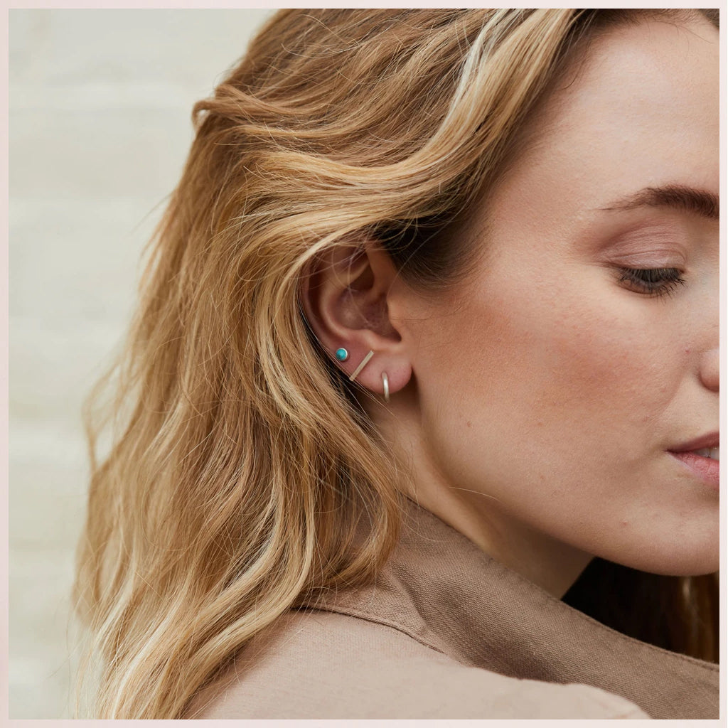 A woman with wavy blonde hair wears Scout Jewelry's Scarlett Stud Trio Turquoise Silver—featuring a semi-precious turquoise stud and a thin silver hoop. She is in a beige top, facing sideways, highlighting her hypoallergenic earrings.